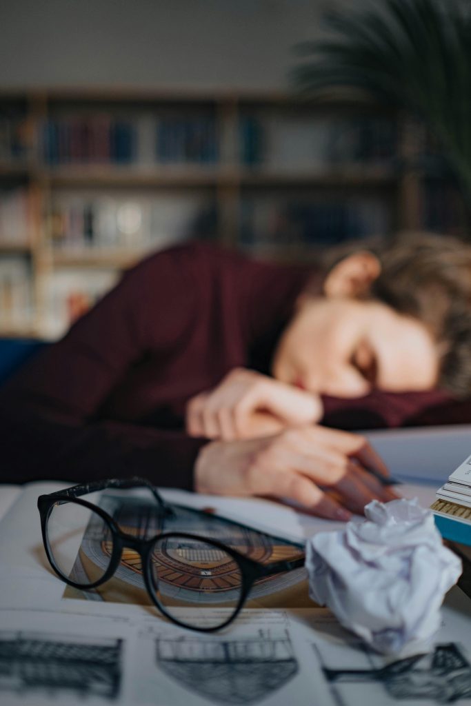 A tired student sleeping on a table surrounded by books, paper, and eyeglasses.